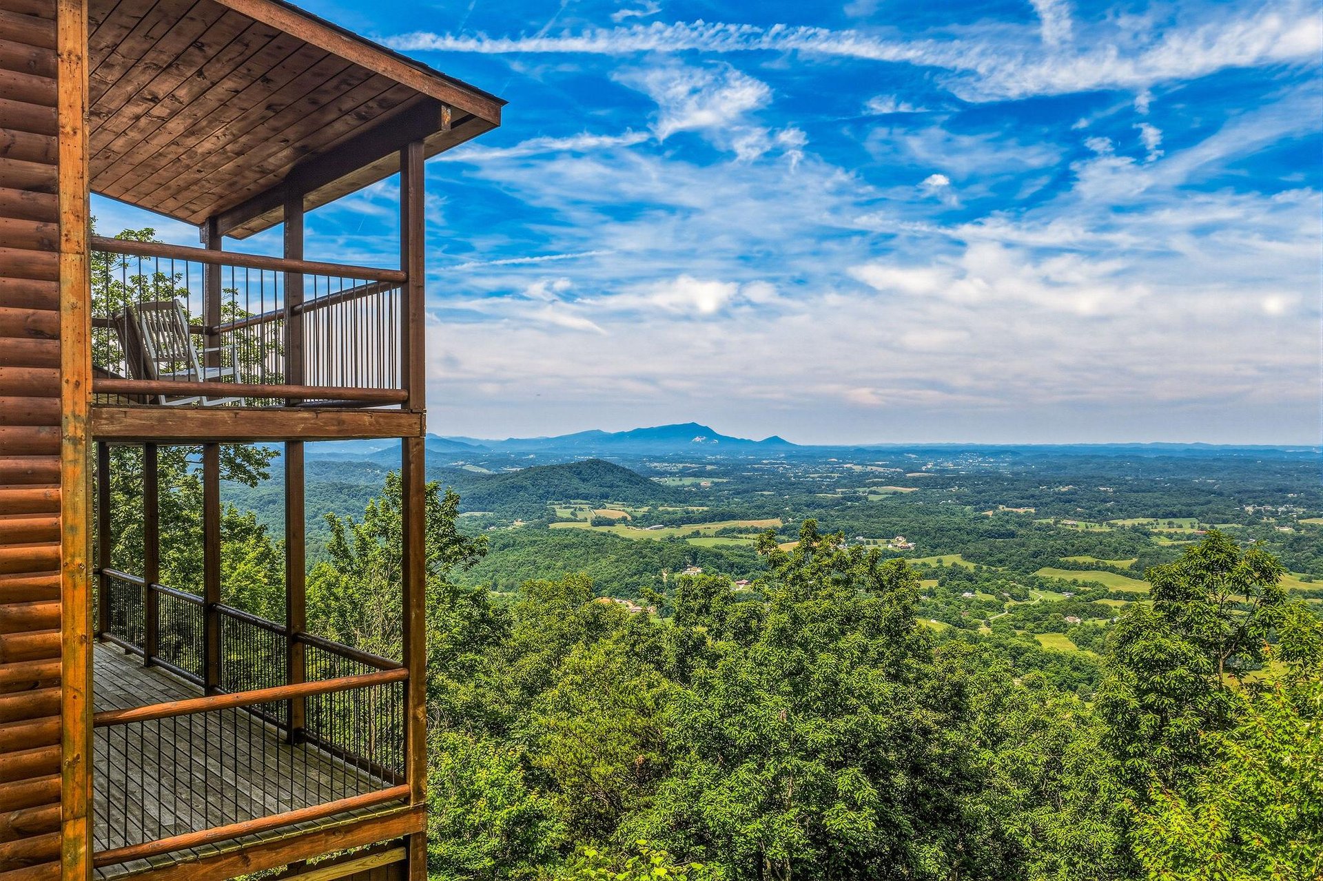 The multilevel balcony structure overlooks expansive valley farmland and distant mountain ridges beneath a dramatic blue sky, offering a scenic backdrop to the propertys elevated forest setting.