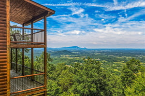 The multilevel balcony structure overlooks expansive valley farmland and distant mountain ridges beneath a dramatic blue sky, offering a scenic backdrop to the propertys elevated forest setting.