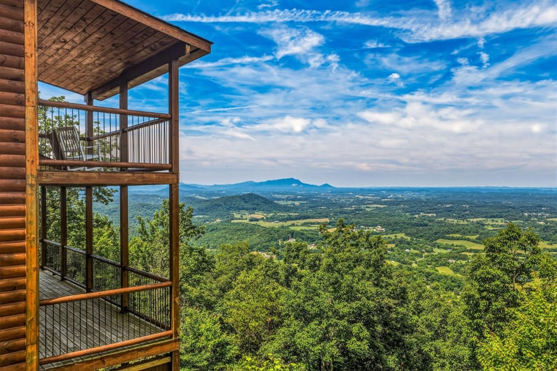 The multilevel balcony structure overlooks expansive valley farmland and distant mountain ridges beneath a dramatic blue sky, offering a scenic backdrop to the propertys elevated forest setting.