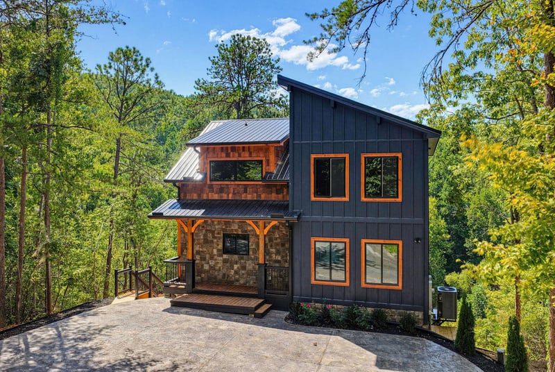 A modern mountain retreat showcasing striking architectural design with bold charcoal siding and warm wood accents, surrounded by lush forest canopy and blue skies overhead.
