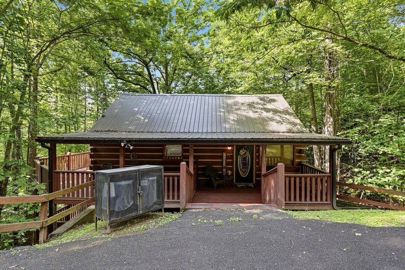 A charming log cabin nestled in lush greenery with a covered porch and convenient parking, offering a peaceful woodland retreat surrounded by towering trees and natural beauty.