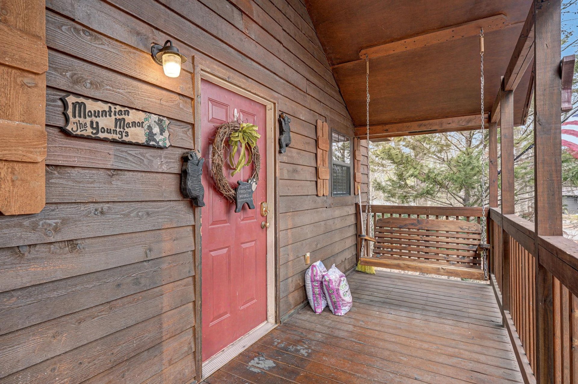 Charming mountain cabin entrance featuring rustic wood siding, welcoming red door with seasonal wreath, and peaceful covered porch with swing for relaxing.