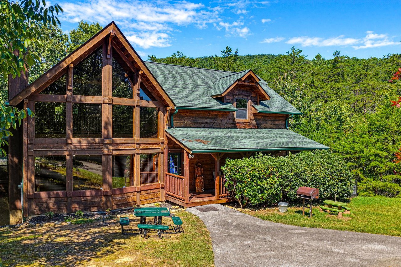 A stunning Aframe log cabin nestled among lush greenery, offering rustic mountain charm and a peaceful wooded setting. The spacious driveway and picnic area set the scene for your arrival.