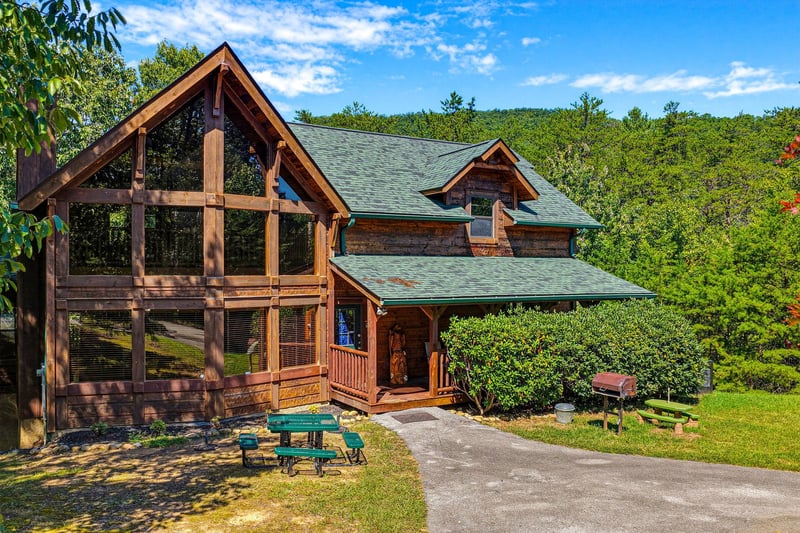 A stunning Aframe log cabin nestled among lush greenery, offering rustic mountain charm and a peaceful wooded setting. The spacious driveway and picnic area set the scene for your arrival.