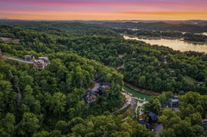 Aerial view captures the property nestled among rolling forests and lakefront beauty, with a stunning sunset painting the sky in vibrant hues over the surrounding landscape.