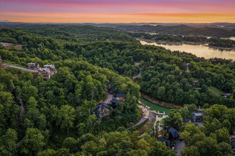 Aerial view captures the property nestled among rolling forests and lakefront beauty, with a stunning sunset painting the sky in vibrant hues over the surrounding landscape.