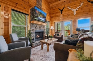 Cozy up in this inviting living room with warm wood walls, a crackling fireplace, and plenty of natural light streaming through windows that frame stunning mountain views beyond.