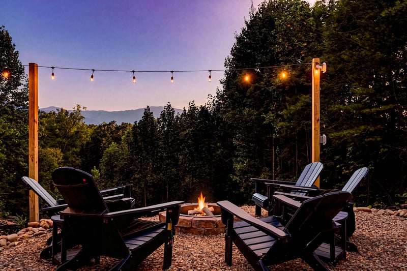 A crackling fire pit surrounded by Adirondack chairs sets the scene for starlit evenings beneath string lights, with mountain ridges fading into twilight just beyond the trees.