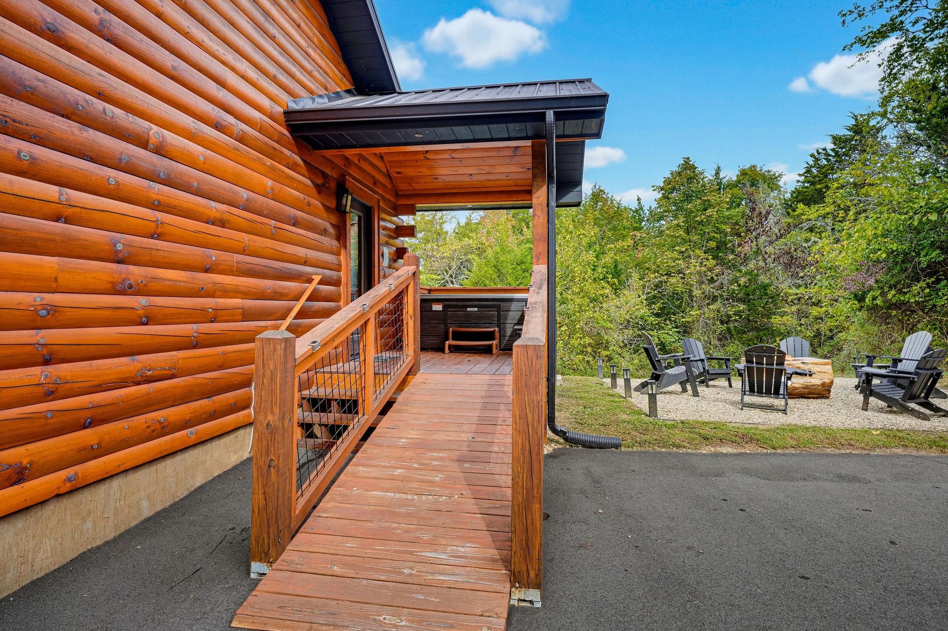 The property features a charming log cabin exterior surrounded by lush greenery, with a private deck and hot tub for your relaxation. Unwind in the fire pit seating area under open skies.