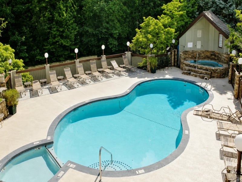 Aerial view of the propertys pool area and surrounding landscape. The pool deck is nestled among lush trees and features a separate hot tub.
