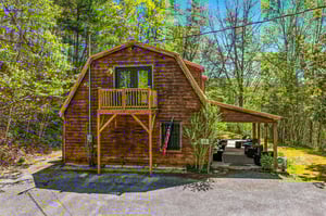 Charming rustic cabin nestled among lush spring foliage with a welcoming balcony and covered parking area.