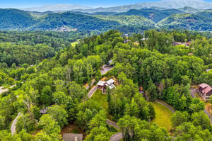 Aerial perspective of the property nestled in a forested mountain valley, showcasing the natural landscape and surrounding residential area with distant mountain vistas.