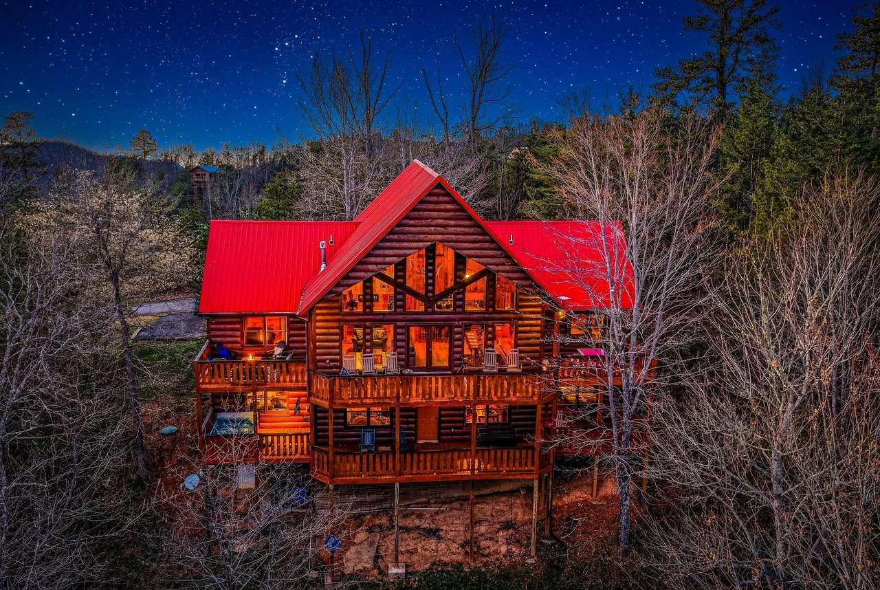 Stunning log cabin retreat glows warmly against a starlit mountain sky, surrounded by peaceful forest landscape.