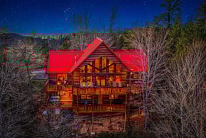 Stunning log cabin retreat glows warmly against a starlit mountain sky, surrounded by peaceful forest landscape.