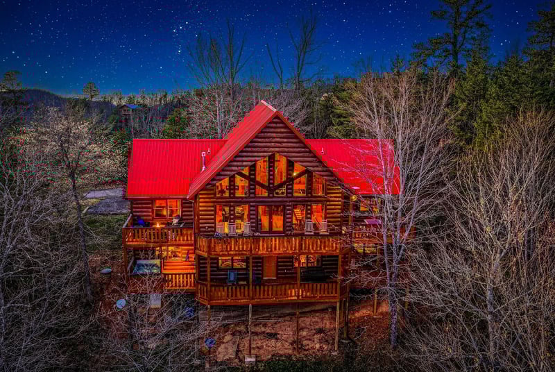 Stunning log cabin retreat glows warmly against a starlit mountain sky, surrounded by peaceful forest landscape.