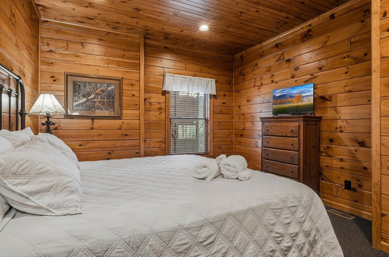 Sink into this cozy cabin bedroom where warm wood paneling wraps around you, fresh towels await your arrival, and a gentle mountain breeze drifts through the window above your plush bed.