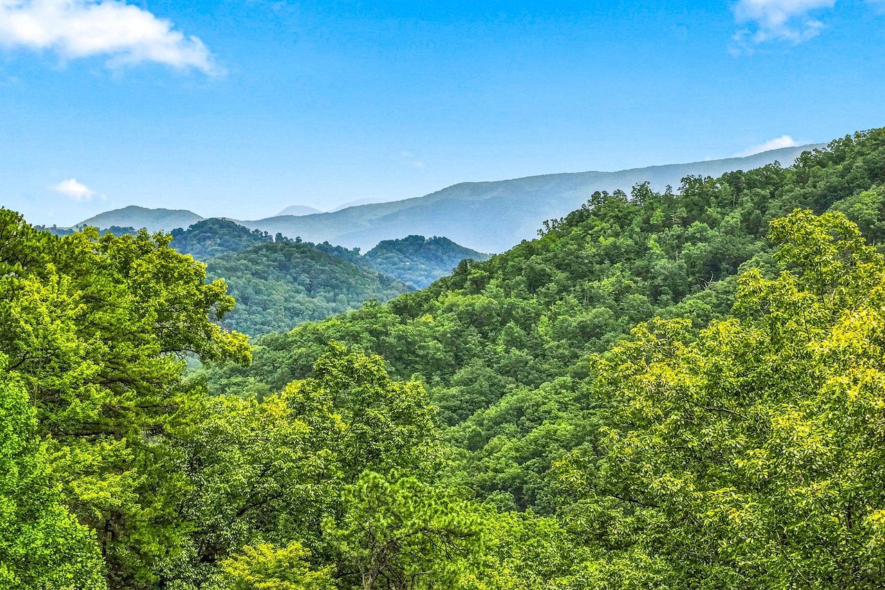 A scenic mountain landscape captured from the property, with rolling forested ridges stretching into the distance beneath a bright blue sky and gentle clouds.