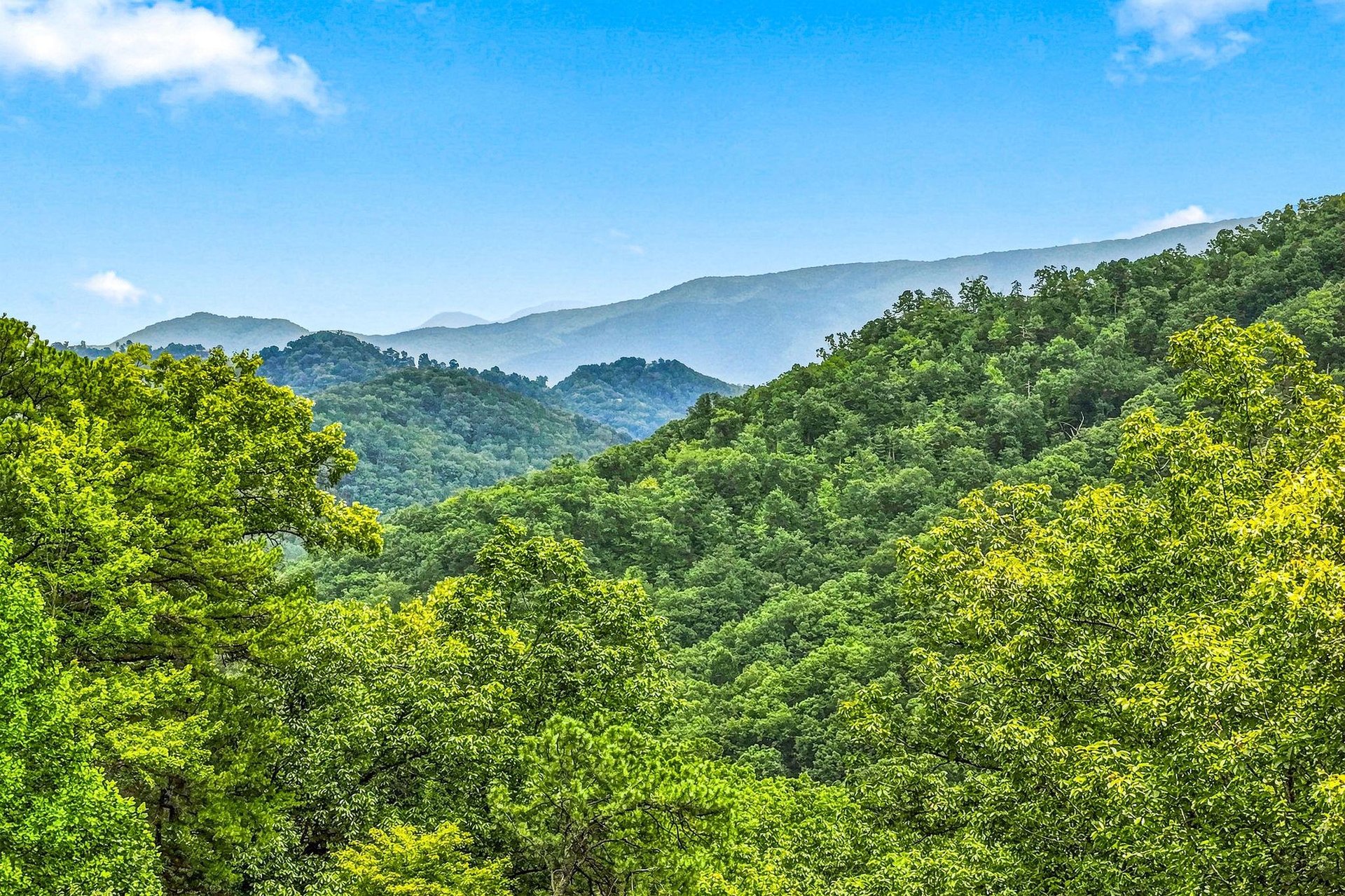 A scenic mountain landscape captured from the property, with rolling forested ridges stretching into the distance beneath a bright blue sky and gentle clouds.