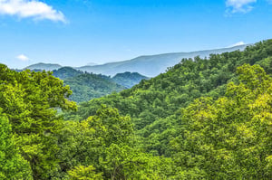 A scenic mountain landscape captured from the property, with rolling forested ridges stretching into the distance beneath a bright blue sky and gentle clouds.