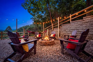 Gather around the glowing fire pit as twilight settles in, sinking into cozy Adirondack chairs with plush cushions beneath strung café lights and a canopy of trees overhead.