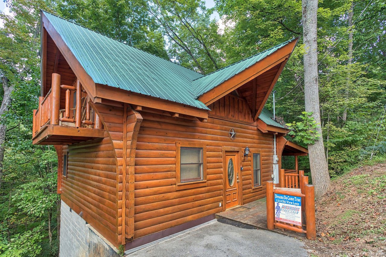 Charming log cabin with green metal roof nestled among towering trees, featuring a welcoming entrance and rustic mountain architecture.
