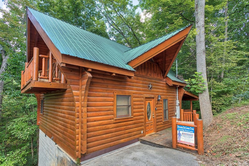 Charming log cabin with green metal roof nestled among towering trees, featuring a welcoming entrance and rustic mountain architecture.