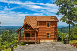 Charming log cabin perched on a mountainside with breathtaking valley views, featuring a welcoming covered porch and surrounded by lush forest greenery.