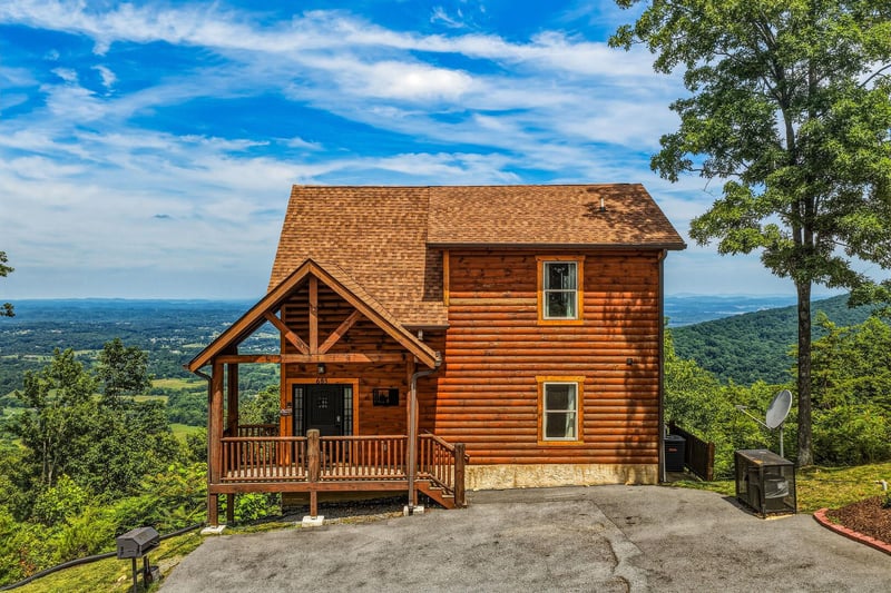 Charming log cabin perched on a mountainside with breathtaking valley views, featuring a welcoming covered porch and surrounded by lush forest greenery.