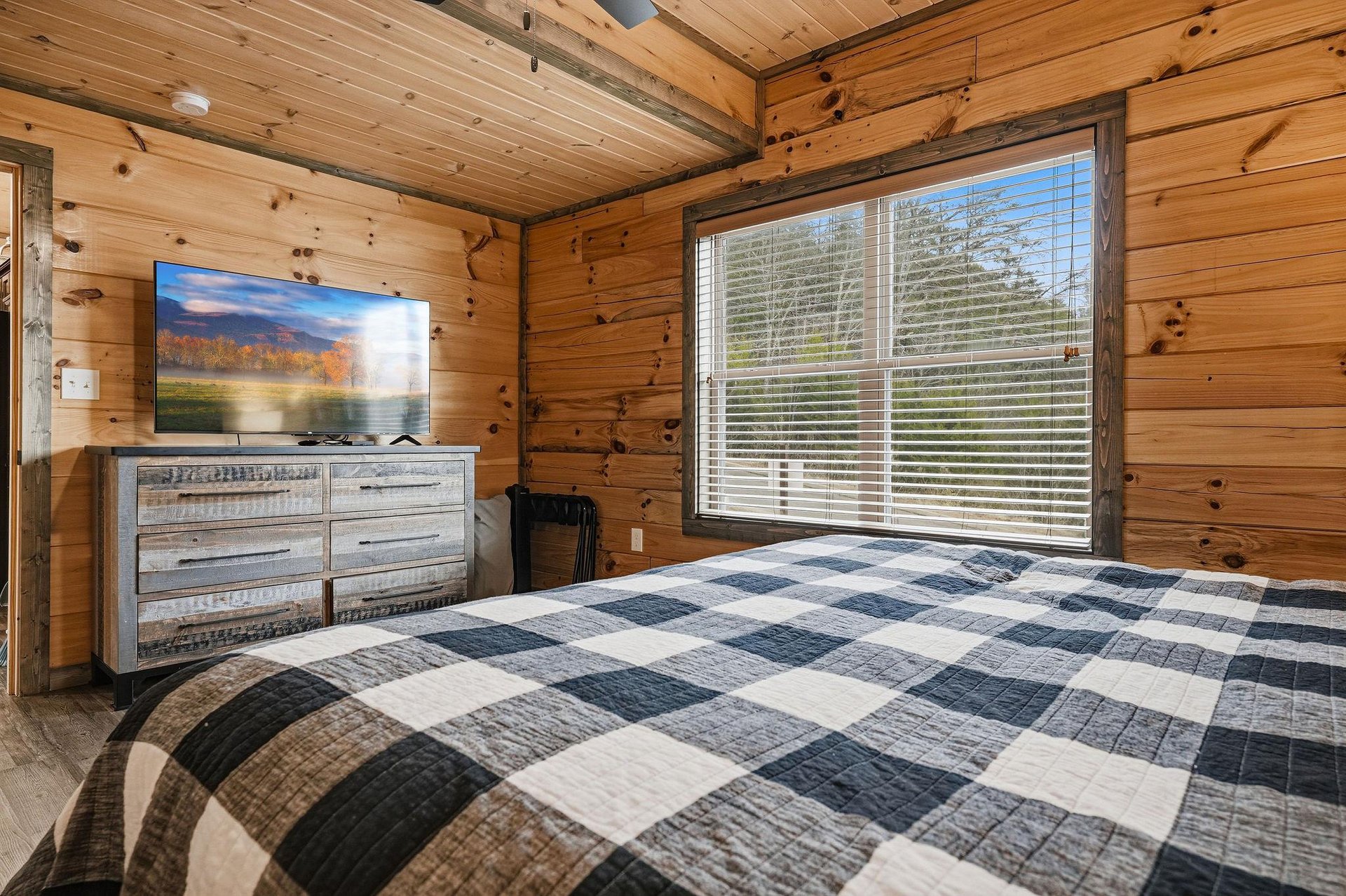Cozy log cabin bedroom with rustic wood walls, flatscreen TV, and large window overlooking peaceful trees for your mountain retreat.
