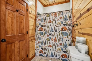 Cozy log cabin bathroom with whimsical campingthemed shower curtain featuring tents, wildlife, and forest scenes.