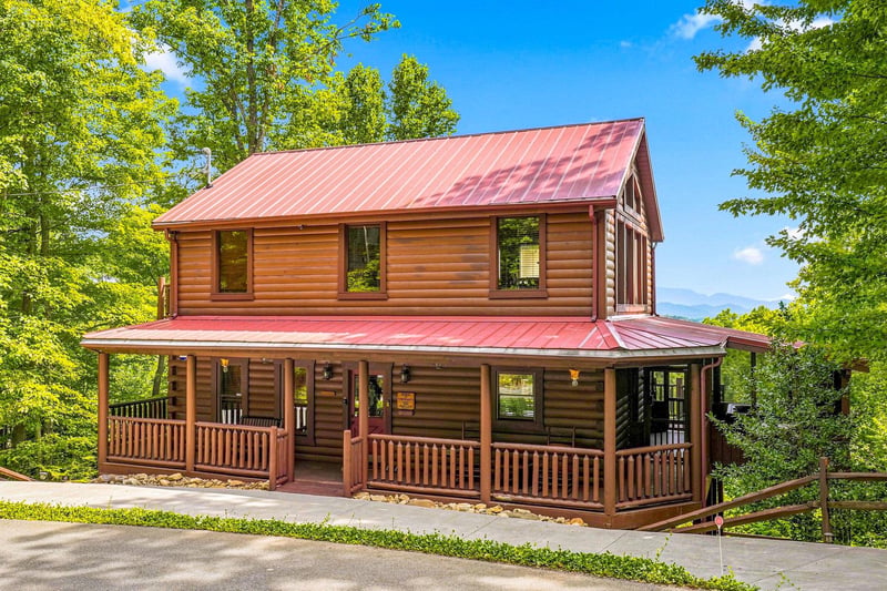 A charming log cabin with a wraparound porch nestled in lush greenery, offering a peaceful mountain escape surrounded by nature.