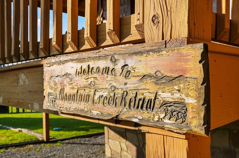 A rustic wooden sign marks the entrance to Mountain Creek Retreat, nestled beneath the propertys wooden deck railing with glimpses of natural surroundings beyond.