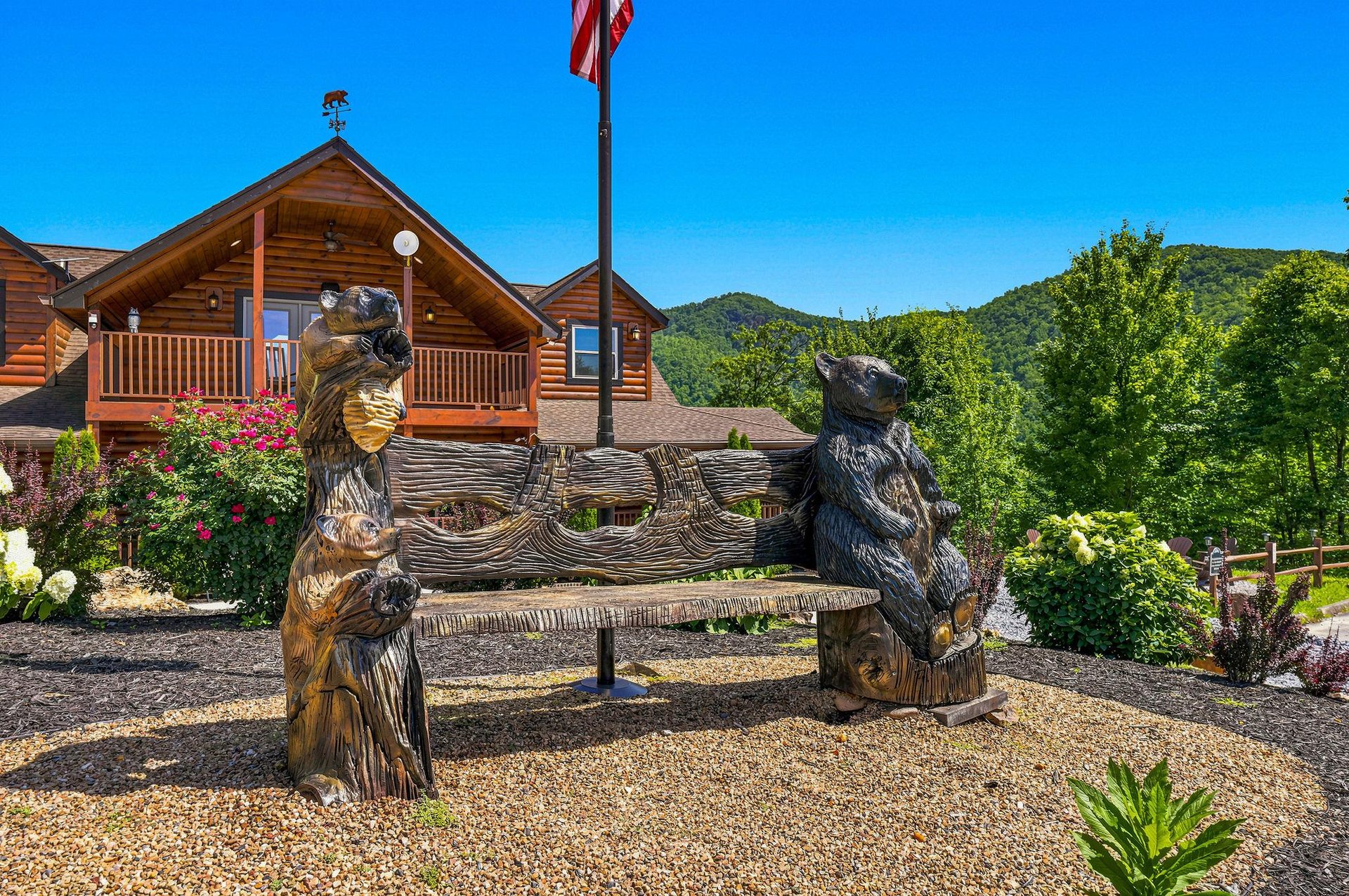 Rustic log cabin nestled in the mountains with decorative carved bear sculptures framing the welcoming entrance area and lush surrounding greenery.