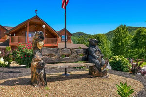 Rustic log cabin nestled in the mountains with decorative carved bear sculptures framing the welcoming entrance area and lush surrounding greenery.