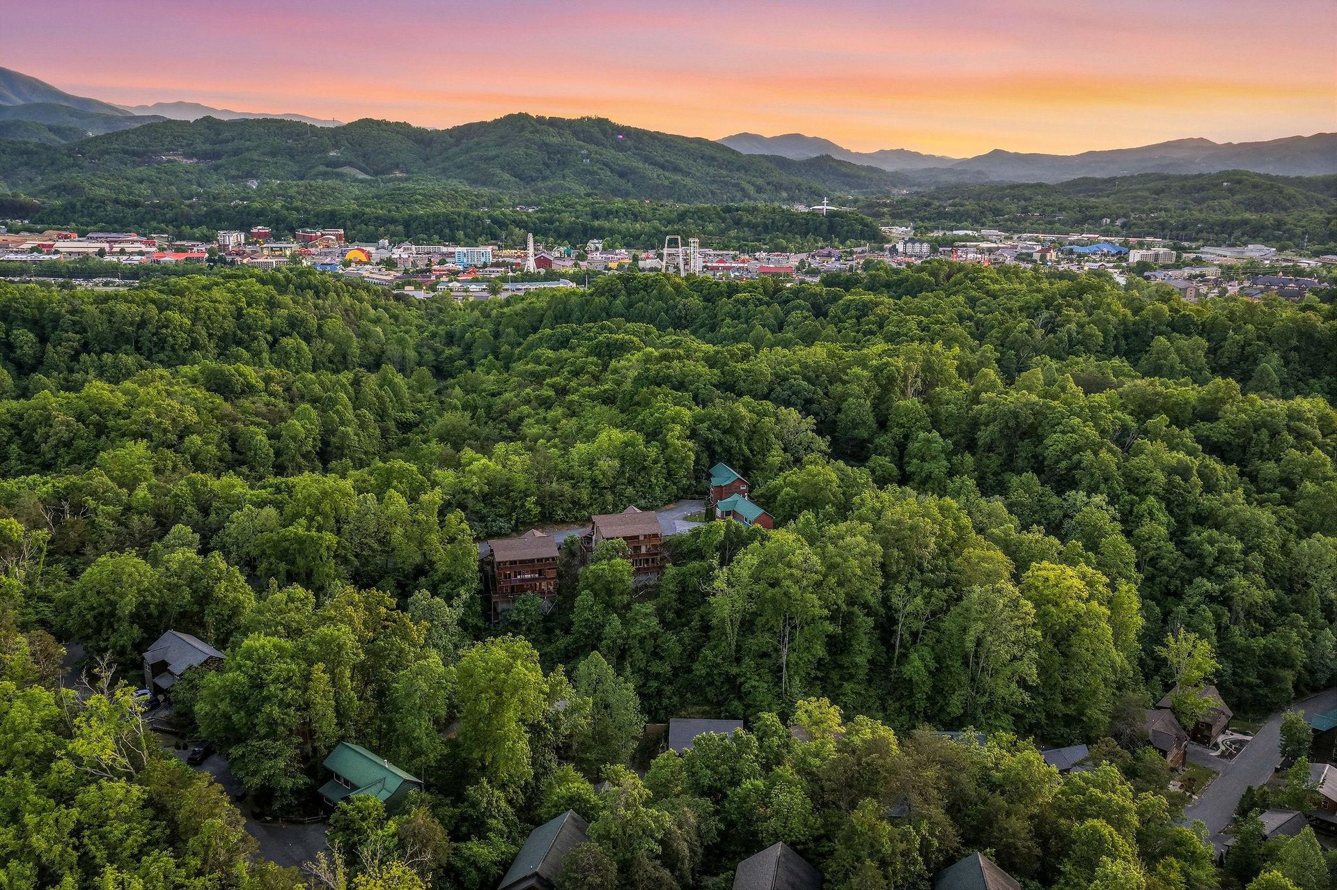 Aerial view captures the property nestled among lush forest with colorful sunset over mountain landscape and nearby town.