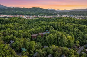 Aerial view captures the property nestled among lush forest with colorful sunset over mountain landscape and nearby town.