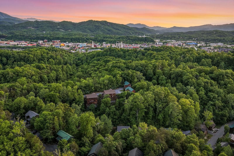 Aerial view captures the property nestled among lush forest with colorful sunset over mountain landscape and nearby town.