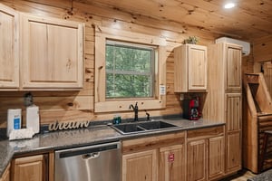 Step into your rustic cabin kitchen with warm pine cabinetry, granite counters, and modern appliances. Natural light streams through the window above the farmhouse sink as greenery peeks in from outsi