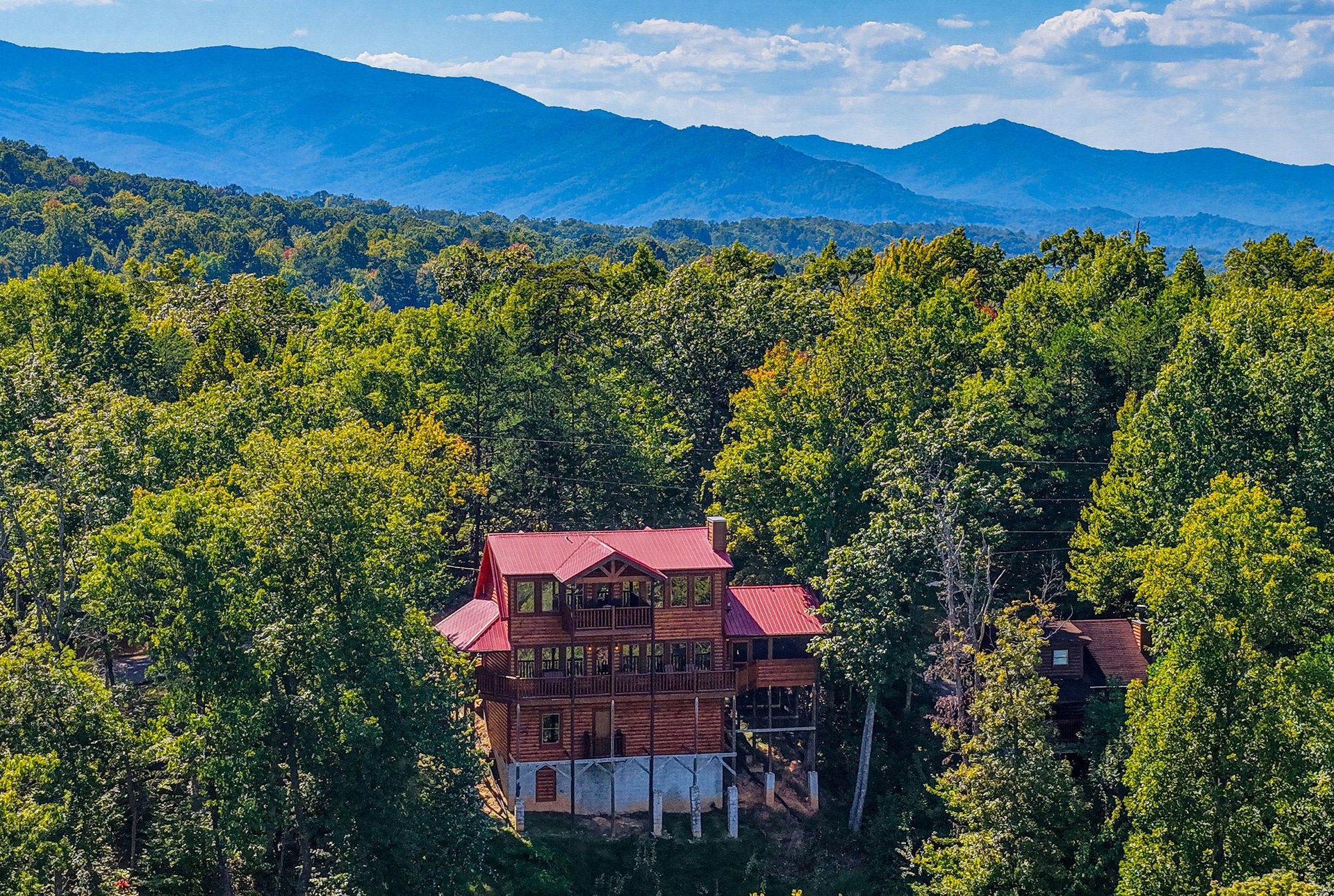 A striking log cabin with a red metal roof nestled among lush trees, with the majestic Blue Ridge Mountains rising in the background under a bright sky.