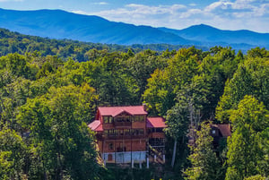 A striking log cabin with a red metal roof nestled among lush trees, with the majestic Blue Ridge Mountains rising in the background under a bright sky.