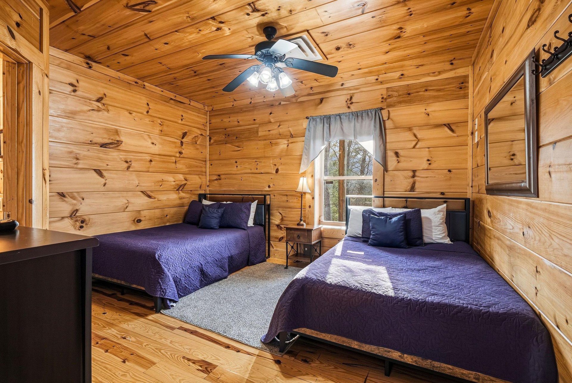 Cozy cabin bedroom with twin beds wrapped in soft purple linens, where rustic wood walls create your peaceful mountain retreat.