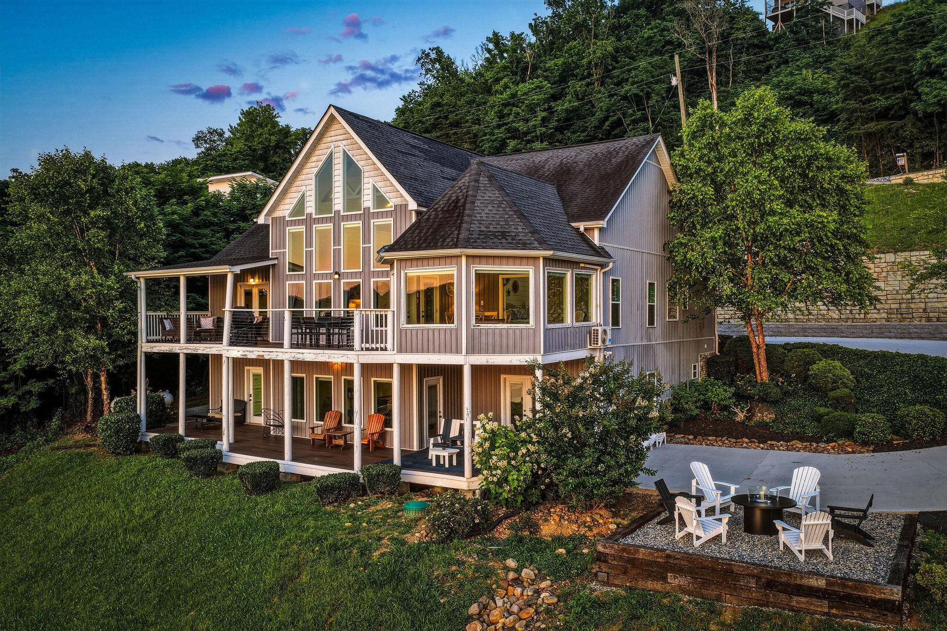 This stunning lakeside retreat features expansive windows, multilevel covered porches, and a private outdoor fire pit area—all nestled among lush greenery and scenic hillside surroundings.