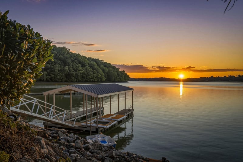 A tranquil lakeside setting features a covered boat dock against a stunning sunset, surrounded by lush forests and calm waters reflecting the evening sky.