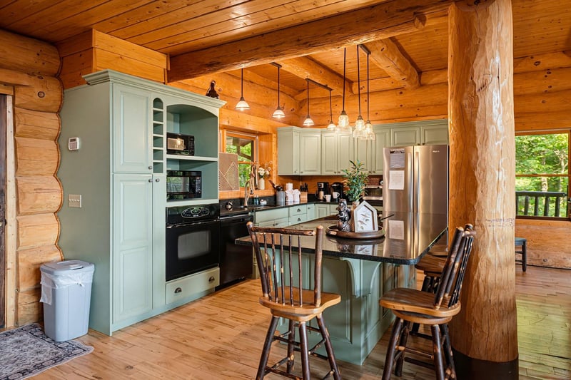 Gather around the rusticmeetscharming kitchen, where vaulted wood ceilings and soft green cabinetry create a cozy, cabin retreat feel. Pendant lights glow above the breakfast bar, inviting you to si