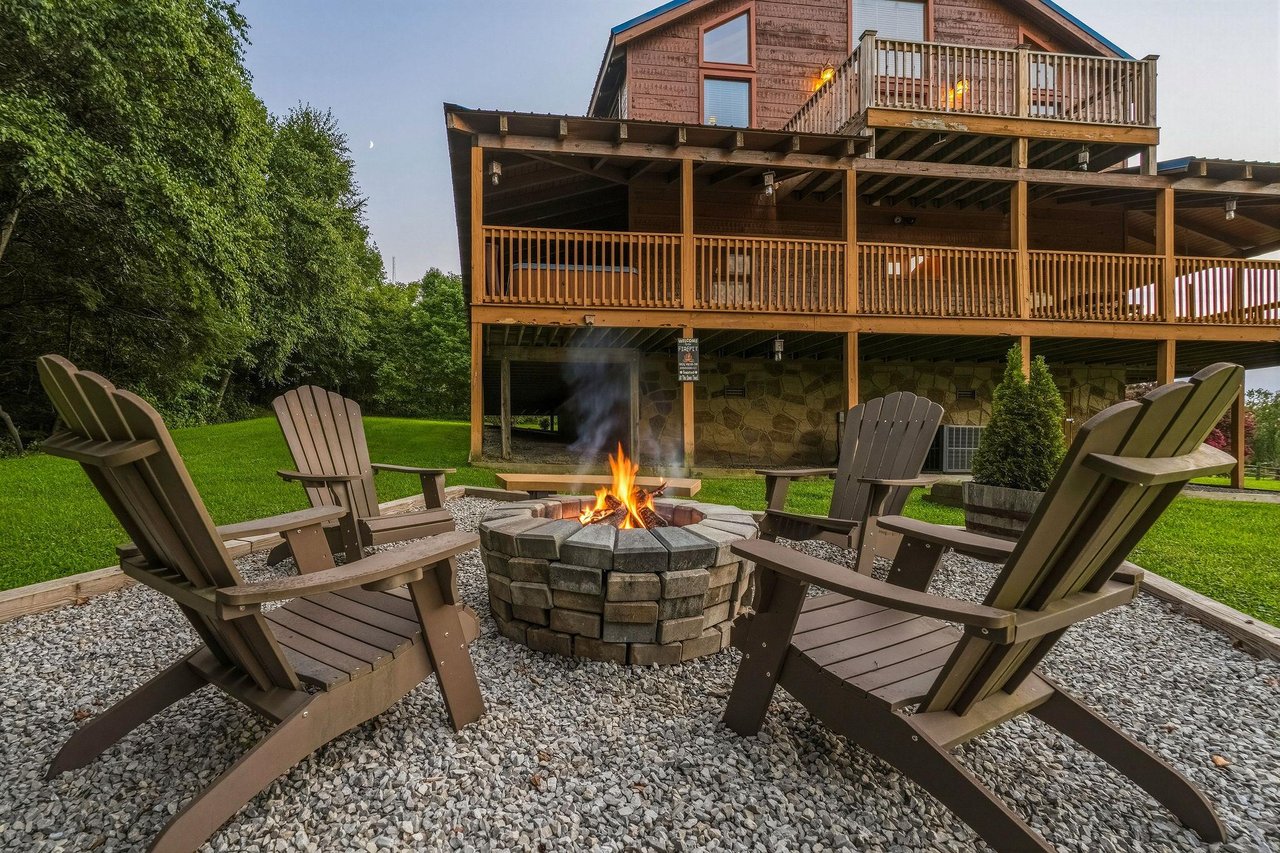 This mountain lodge features expansive wooden decks on multiple levels, surrounded by lush green lawn and mature trees. A stone fire pit with Adirondack chairs creates the perfect gathering spot for e