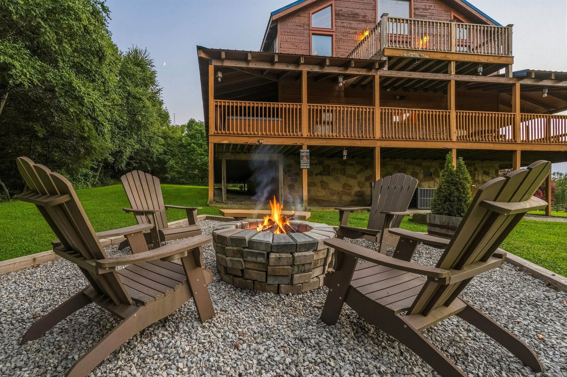 This mountain lodge features expansive wooden decks on multiple levels, surrounded by lush green lawn and mature trees. A stone fire pit with Adirondack chairs creates the perfect gathering spot for e