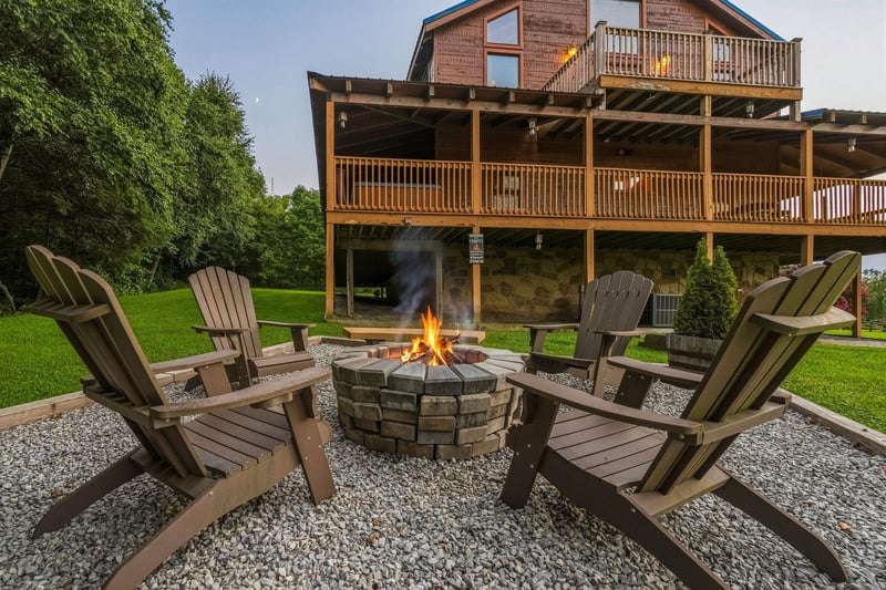 This mountain lodge features expansive wooden decks on multiple levels, surrounded by lush green lawn and mature trees. A stone fire pit with Adirondack chairs creates the perfect gathering spot for e