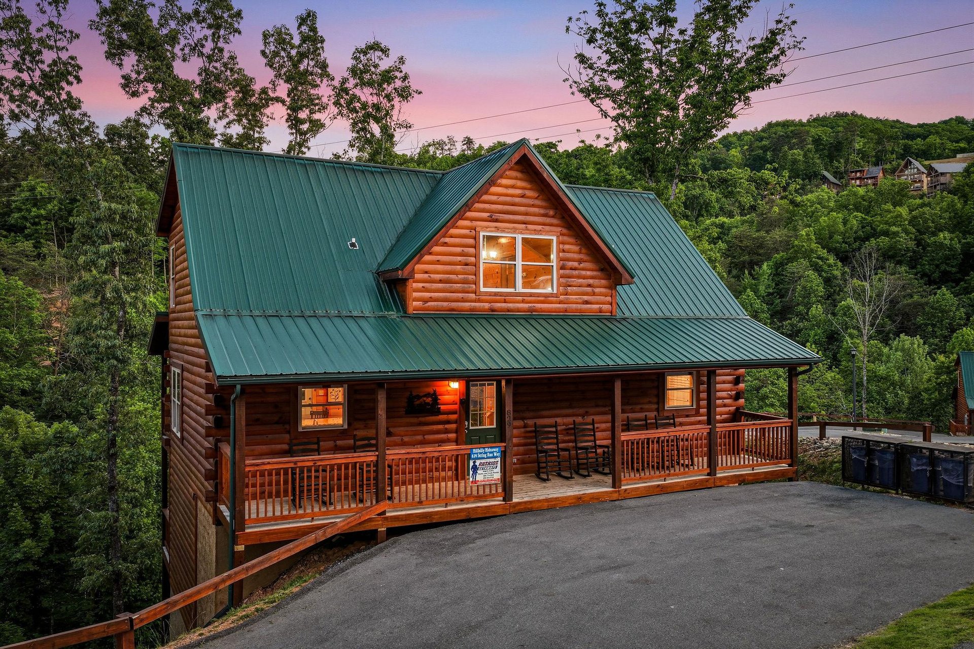 A welcoming log cabin with green metal roof and wraparound porch sits nestled among trees, framed by stunning pink and purple twilight skies in a peaceful mountain setting.