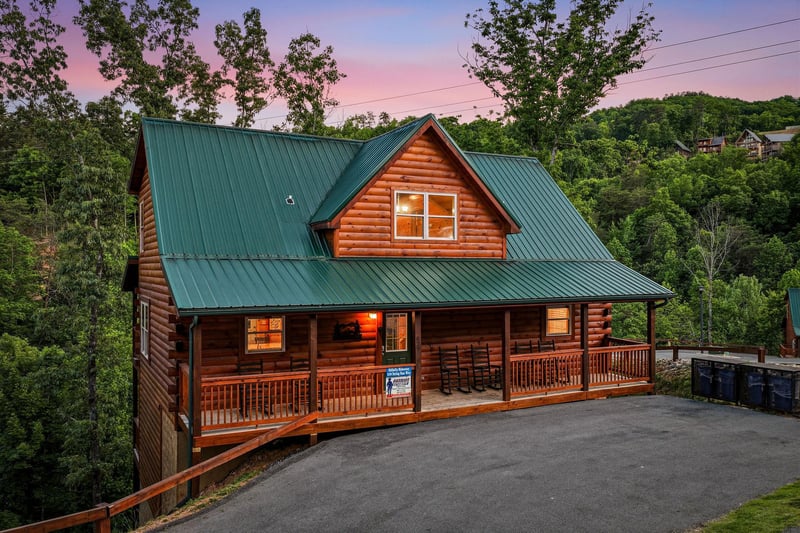 A welcoming log cabin with green metal roof and wraparound porch sits nestled among trees, framed by stunning pink and purple twilight skies in a peaceful mountain setting.