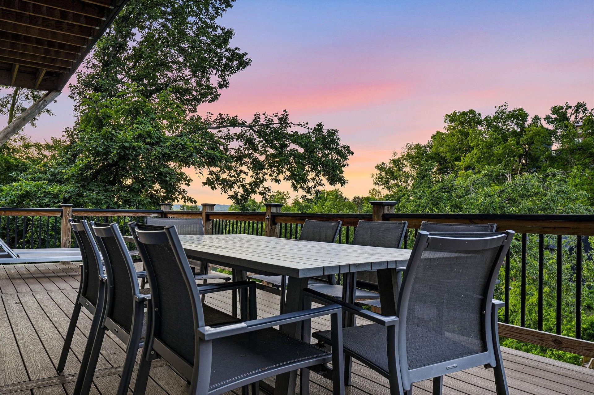 The propertys spacious deck provides a peaceful outdoor dining setting surrounded by mature trees and natural greenery, ideal for evening gatherings under colorful skies.